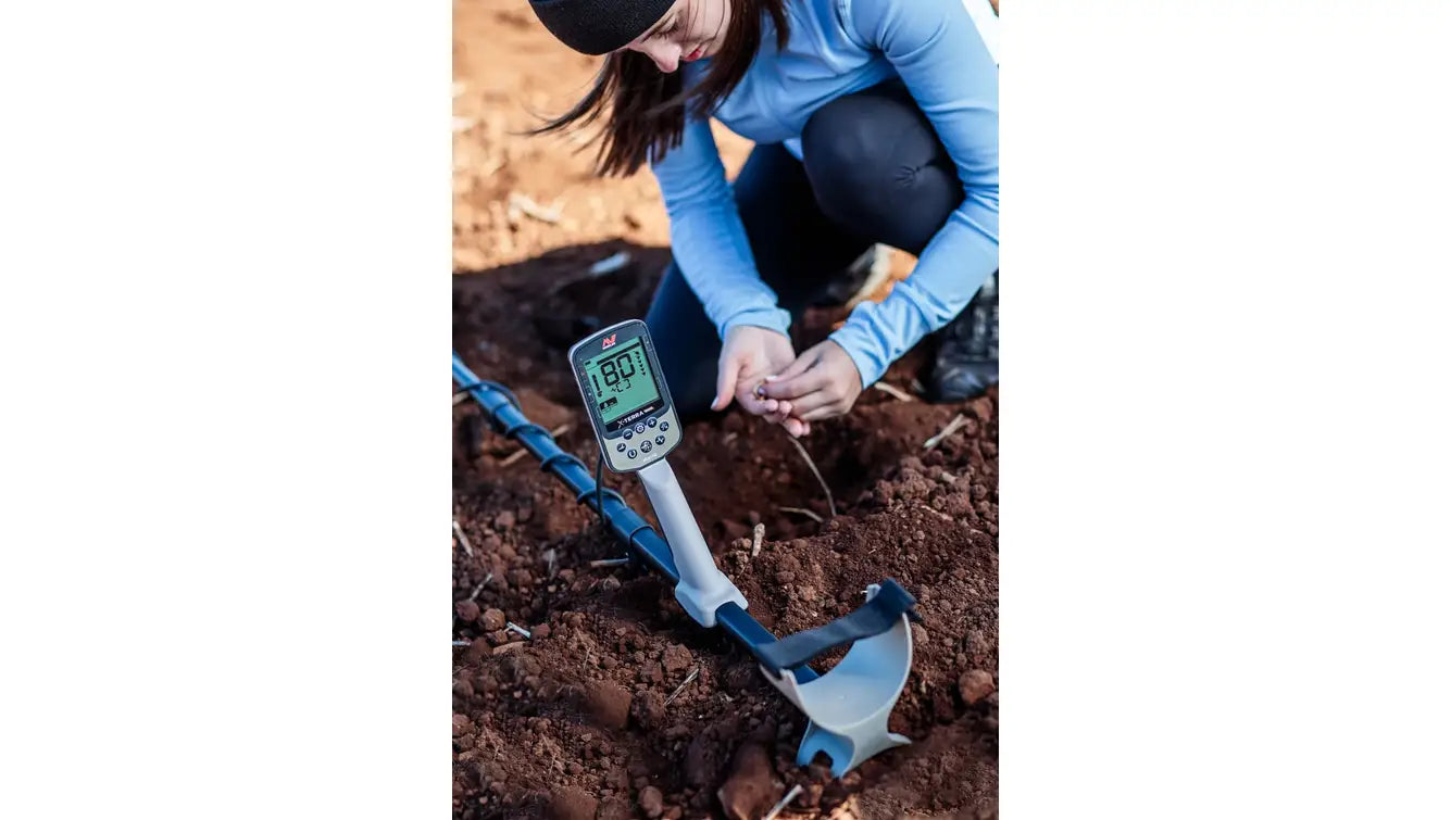 Detectorist kneeling in reddish soil using a Minelab X-Terra Elite to examine a recovered coin